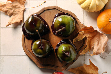 Wooden board of tasty caramel apples for Halloween and autumn leaves on white tile background