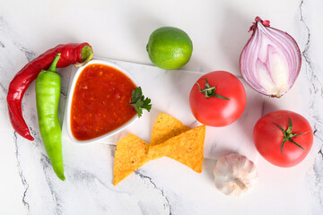 Bowl with tasty salsa sauce, nachos and ingredients on white marble background