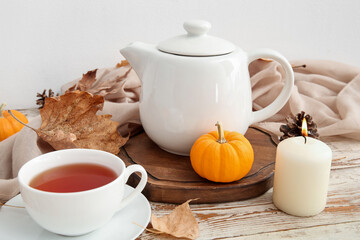 Tea pot, cup of hot drink, pumpkins, burning candle and autumn leaves on table near white wall