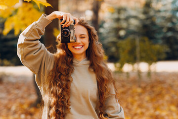 Smiling young woman photographer with the photo camera shooting the forest trees with the yellow leaves at sunset - obrazy, fototapety, plakaty