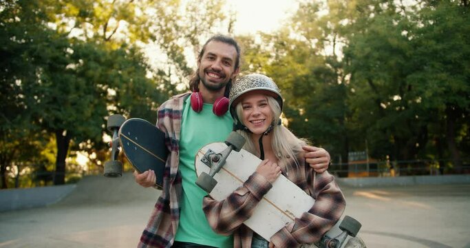 A blonde girl in a leopard print helmet and a plaid shirt stands with a brunette guy in a Green T-shirt and red headphones in a skatepark. A guy and a girl hold a white and black skateboard in their