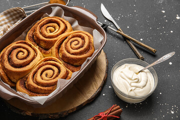 Baking dish with tasty cinnamon rolls and cream on black background