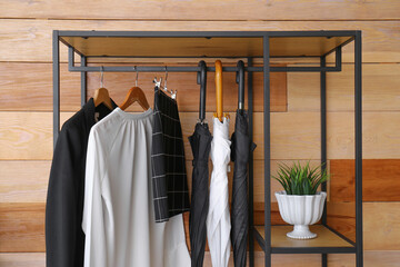 Rack with clothes, umbrellas and plant near wooden wall, closeup