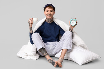 Tattooed young man in pajamas with bedding, alarm clock and cup of coffee sitting on grey background