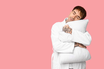 Tattooed young man in pajamas with pillow on pink background