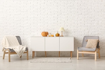 Cabinet with pumpkins, candles and armchairs near white brick wall