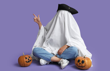 Woman in Halloween costume of ghost with pumpkins sitting and pointing at something on lilac background