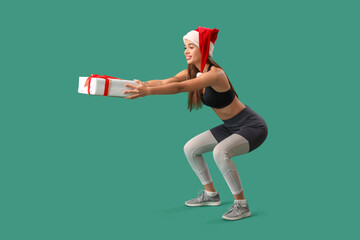 Sporty young woman in Santa hat with Christmas gift training on green background