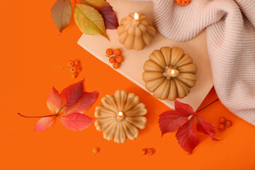 Beautiful composition with burning candles in shape of pumpkin, book and autumn leaves on orange background