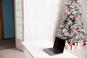 Laptop on table and decorated Christmas tree with gifts in white room interior