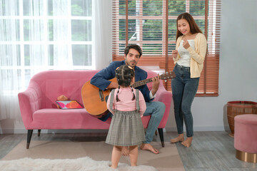 father sitting on sofa playing guitar,a little daughter looking at him while mother standing,sing a song,family spend time together in the living room,concept family,relationship,the love of a family