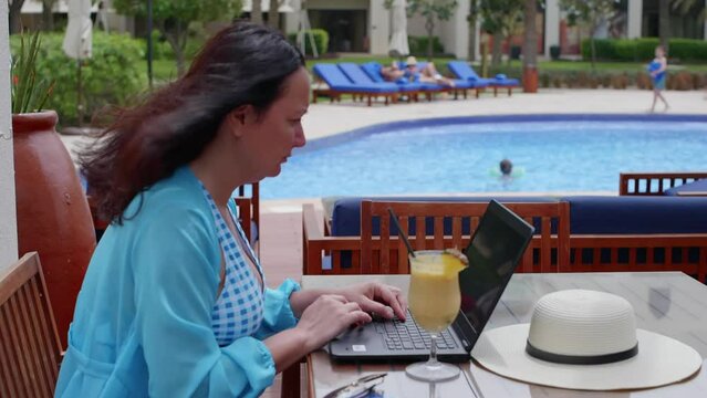 Woman freelancer in swimsuit working on laptop on vacation sitting in cafe in hotel near pool. Brunette middle aged female doing jon typing looking at screen. Remote, distant, freelance work concept.
