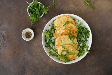 Zucchini pancakes with arugula in a white plate on a brown background. Top view.