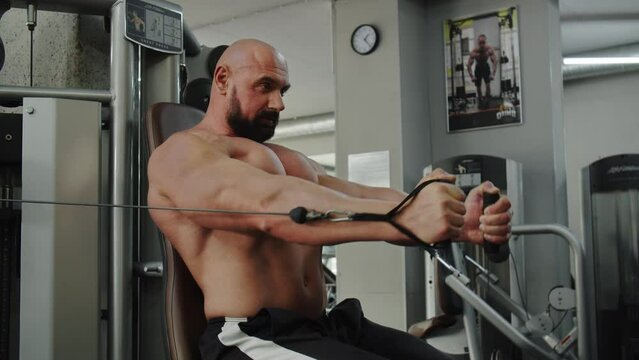 Man bodybuilder in the gym sits on exercise machine with cable. The athlete is flexing, connects and separates his arms. Showcasting of training the muscles of chest.