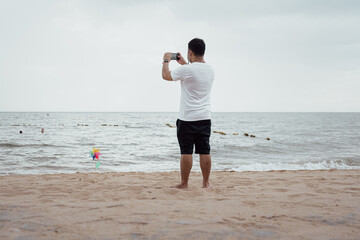 An Asian relaxing young man standing on the beach and using a smartphone takes a photo on the beach
