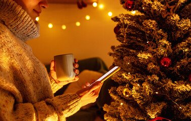 Woman holding cup of hot chocolate and using her smart phone in front of Christmas tree at home. - Powered by Adobe