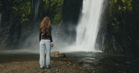 Tourist girl raising arms standing at powerful waterfall Air Terjun Munduk, Indonesia, Bali island. Woman posing, back view. Beautiful nature landscape. Travel, tourism, holiday