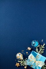 Christmas vertical banner template with glitter blue paper gift box, gold and blue Xmas balls ornaments, fir branches on dark blue background. Flat lay, top view, copy space.