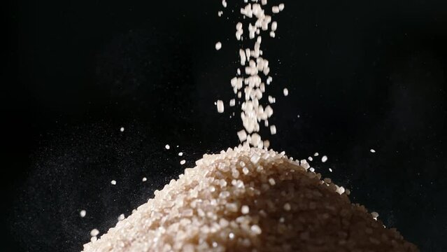 Brown cane sugar is poured into pile. Close up slow motion black background. Protect your health by reducing sugar consumption, as it can lead to obesity diabetes, and other severe ailments. Nutrition