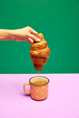 Woman dipping fresh croissant into cup with coffee, latte isolated over pink green background....