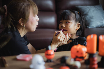 Woman doing halloween make-up for a little cheerful girl in costume while preparing holiday at home