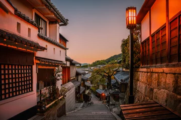 Gordijnen Kyoto City historic old town at sunrise with the warm blue sky in Japan, an empty downhill street with rustic wooden houses, cherry trees, and illuminated street light lamp © Naya Na