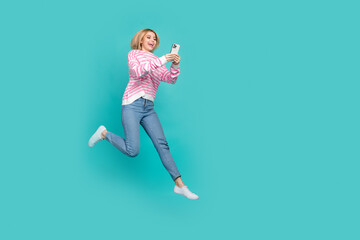 Full length photo of cute excited lady wear pink pullover texting modern device jumping empty space isolated turquoise color background