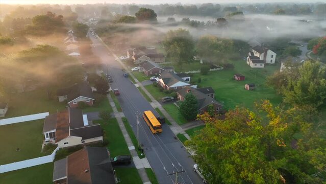 Yellow school bus driving through residential neighborhood at sunrise. Aerial drone shot driving through fog lit by golden hour light. American school transportation in USA.