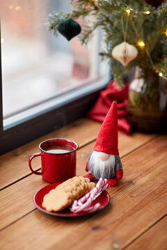 holidays, decoration and celebration concept - close up of christmas gnome, cup of coffee in red mug, ginger cookies and candy canes on window sill at home