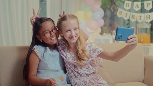 Medium long shot of diverse teen girls taking selfie on smartphone while sitting on couch at birthday party