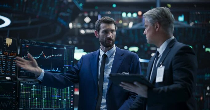 Team of Two Handsome Stock Exchange Brokers Using Tablet Computer, Chatting About the Asset Management Strategy for an International Business Client, Working in a Office Building with Computer Screens