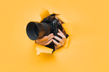 A young paparazzi girl holds a reflex camera and looks through a torn hole in yellow paper. The concept of embarrassment, shame for what he saw. Copy space. - obrazy, fototapety, plakaty