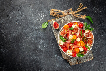 fig salad with mozzarella, prosciutto, fresh basil leaves and tomato on a dark background. top view. place for text