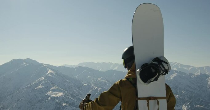 Snowboarder climbed mountain raises hand, celebrating victory, rejoicing at achievement, overcoming difficult way up. Epic mountain landscape of snowy peaks. Back view of snowboarder with snowboard.