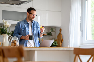 Handsome businessman holding coffee cup and looking at credit card while shopping online in kitchen