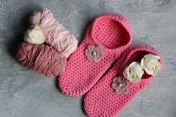 Crochet slippers made of pink cotton tread and balls of yarn on grey background with copy space, closeup photo of hand made footwear. 
