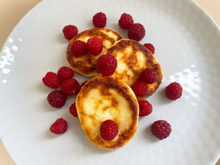 Three fried brown cheesecakes lie in a pile in the center of a gray round plate strewn with small berries of ripe red raspberries on a beige background, close-up, top view. Proper nutrition.