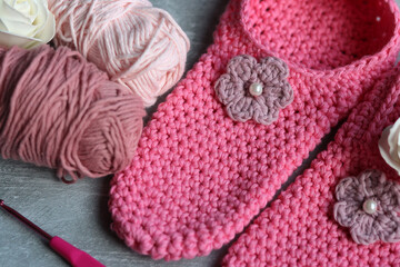 Crochet slippers made of pink cotton tread and balls of yarn on grey background with copy space, closeup photo of hand made footwear. 