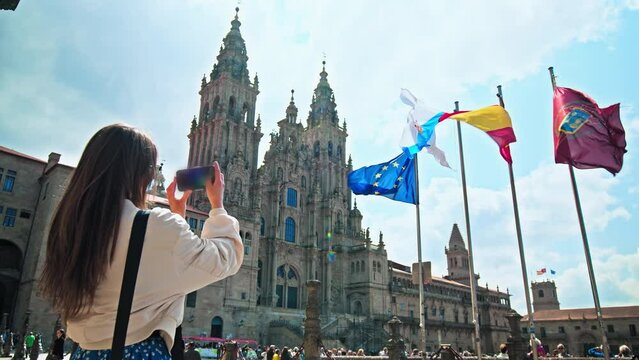 A tourist woman taking a picture of the Cathedral of Santiago de Compostela. A female photographing with a phone flags of Galicia, Spain, Europe on a European historic World Heritage Site
