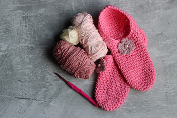 Crochet slippers made of pink cotton tread and balls of yarn on grey background with copy space, closeup photo of hand made footwear. 