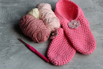 Crochet slippers made of pink cotton tread and balls of yarn on grey background with copy space, closeup photo of hand made footwear. 