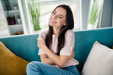 Photo of adorable positive lady sit couch closed eyes beaming smile hands embrace shoulders modern apartment inside