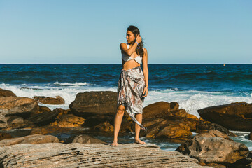 young woman modeling on the beach on the rocks with the sea in the background. dressed in a sarong...