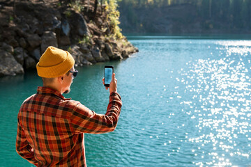 man dressed in plaid shirt and yellow beanie captures the moment on his smartphone by blue serene mountain lake photographing the landscape copy space
