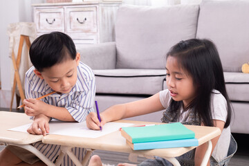 Two Asian siblings boy and girls having fun laughing and drawing cartoons on paper in living room on holiday at home, family relationship childhood lifestyle playing together, school kids free time