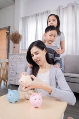 Asian family teaching saving for boy an girl, young mother happy placing coins to a piggy bank and...
