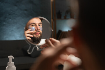 young man in white bathrobe in front of mirror in bathroom putting under eye patches on face
