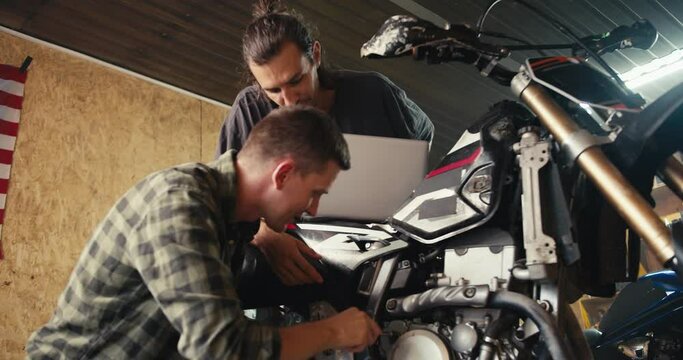 A more experienced mechanic is helping his trainee to properly disassemble moped parts in the workshop. Happy mechanics in their garage workshop inspecting and repairing a motorcycle
