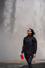 Traveller in black waterproof jacket near big waterfall with rainbow. Tourist girl holding red thermos cup with hot tea in hands. Travel in Iceland. Travel concept. © Iryna