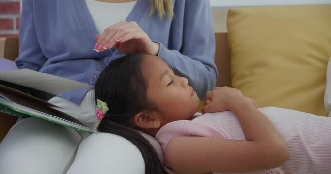 Asian little girl sleeping on mother's lap while listening story book or fairy tales from mother in living room at home. Family relationships concept